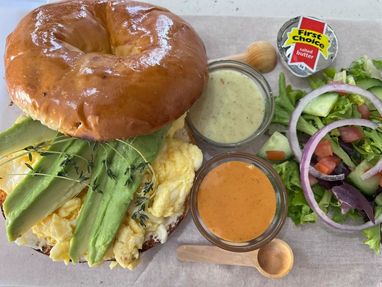 Toasted bagel stacked with scrambled eggs and avocado, served with two house sauces and a side salad