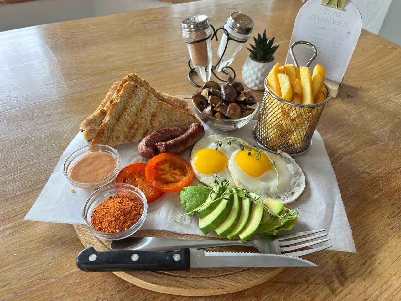 Chew's big breakfast — fried eggs, sausage, avocado fan, grilled tomato, mushrooms, toast and a basket of golden fries on a wooden table